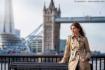 Frau im hellen Trenchcoat am Themse-Ufer vor der Tower Bridge.
