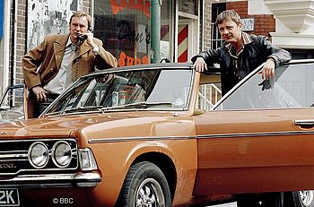 Zwei Männer stehen in den 1970er-Jahren an einem Auto mit geöffneten Türen. Ein Mann telefoniert.