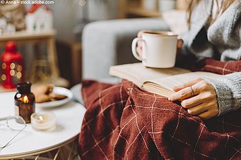 Eine Frau sitzt mit einer Tasse Tee in der Hand und einer Decke auf den Beinen auf einem Sessel und liest ein Buch