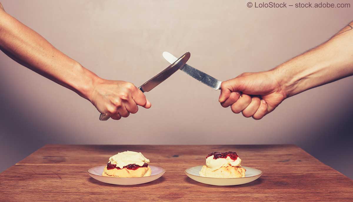 Zwei Hände kreuzen die Messerklingen über einem Tisch, auf dem zwei Teller jeweils mit einem Scone, Clotted Cream und Konfitüre stehen.